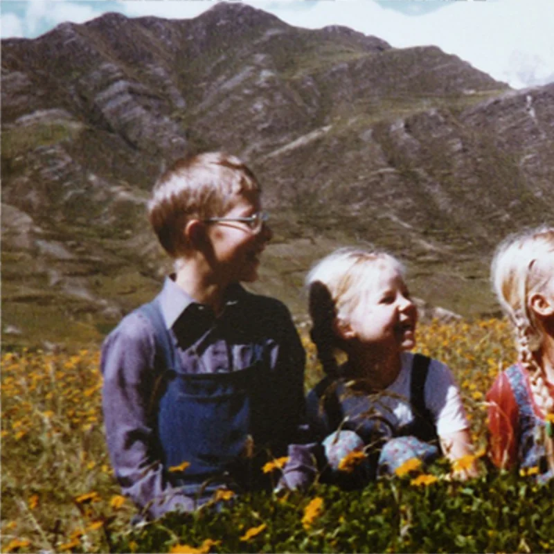 Tres niños sentados entre flores silvestres frente a una montaña rocosa.