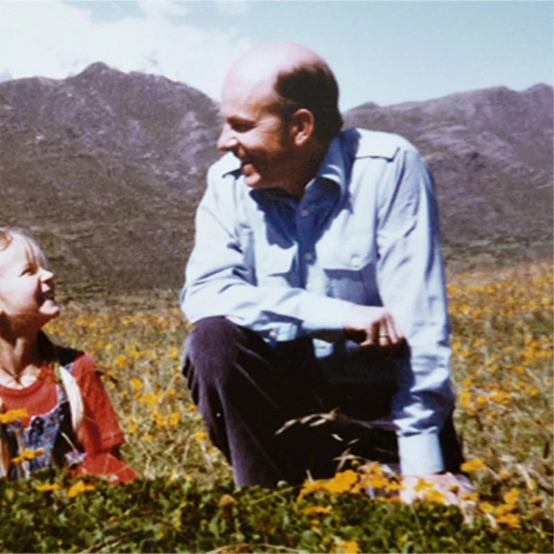 Hombre y niña sentados entre flores silvestres, con montañas al fondo.